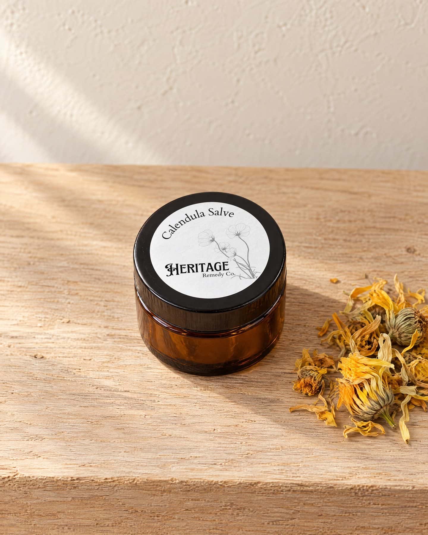 A jar of Calendula Salve, its round label on the lid, on a pale oak shelf with a few dried calendula flowers
