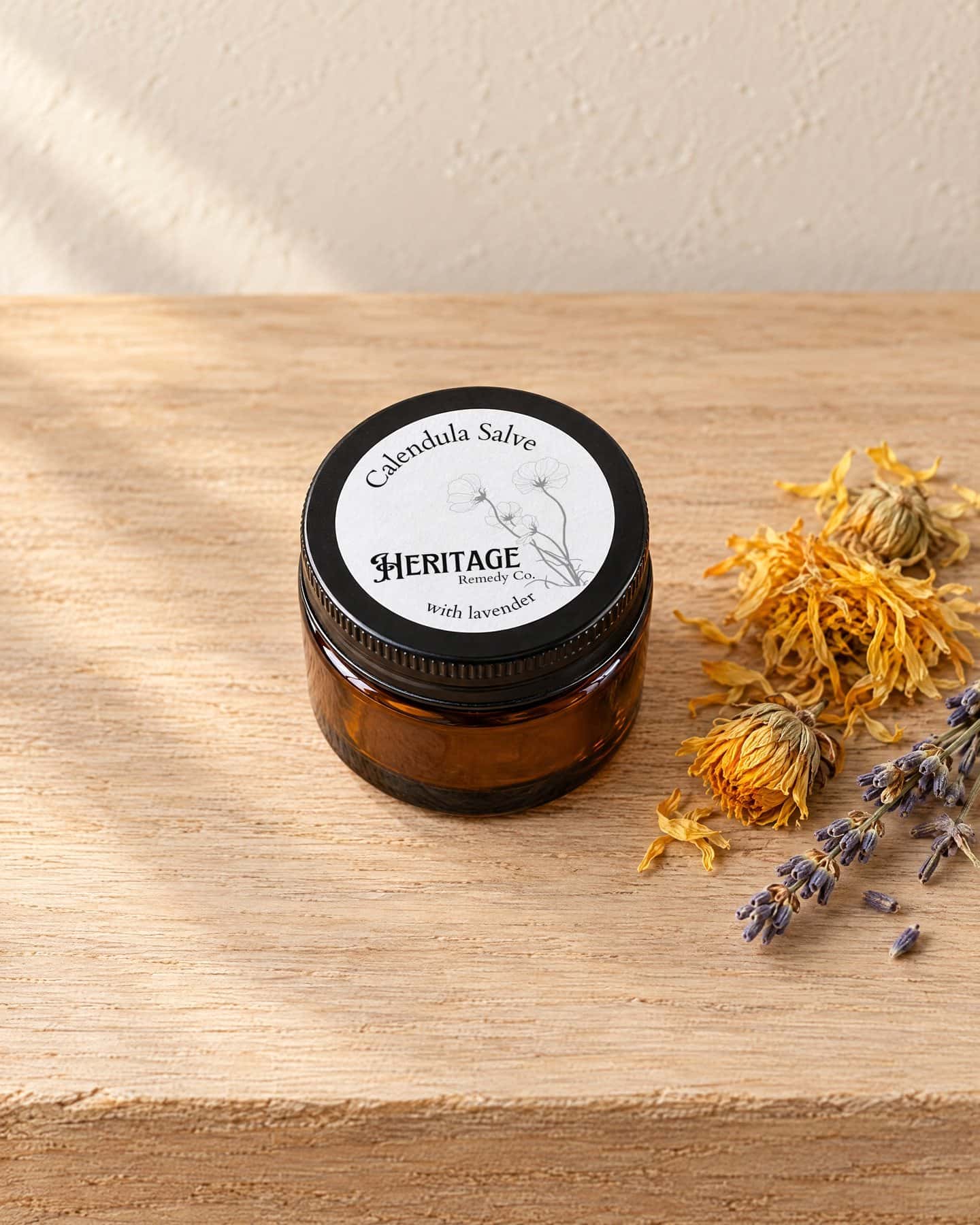 A jar of Calendula with Lavender Salve, its round label on the lid, on a pale oak shelf with a few dried calendula flowers and a sprig of dried lavender