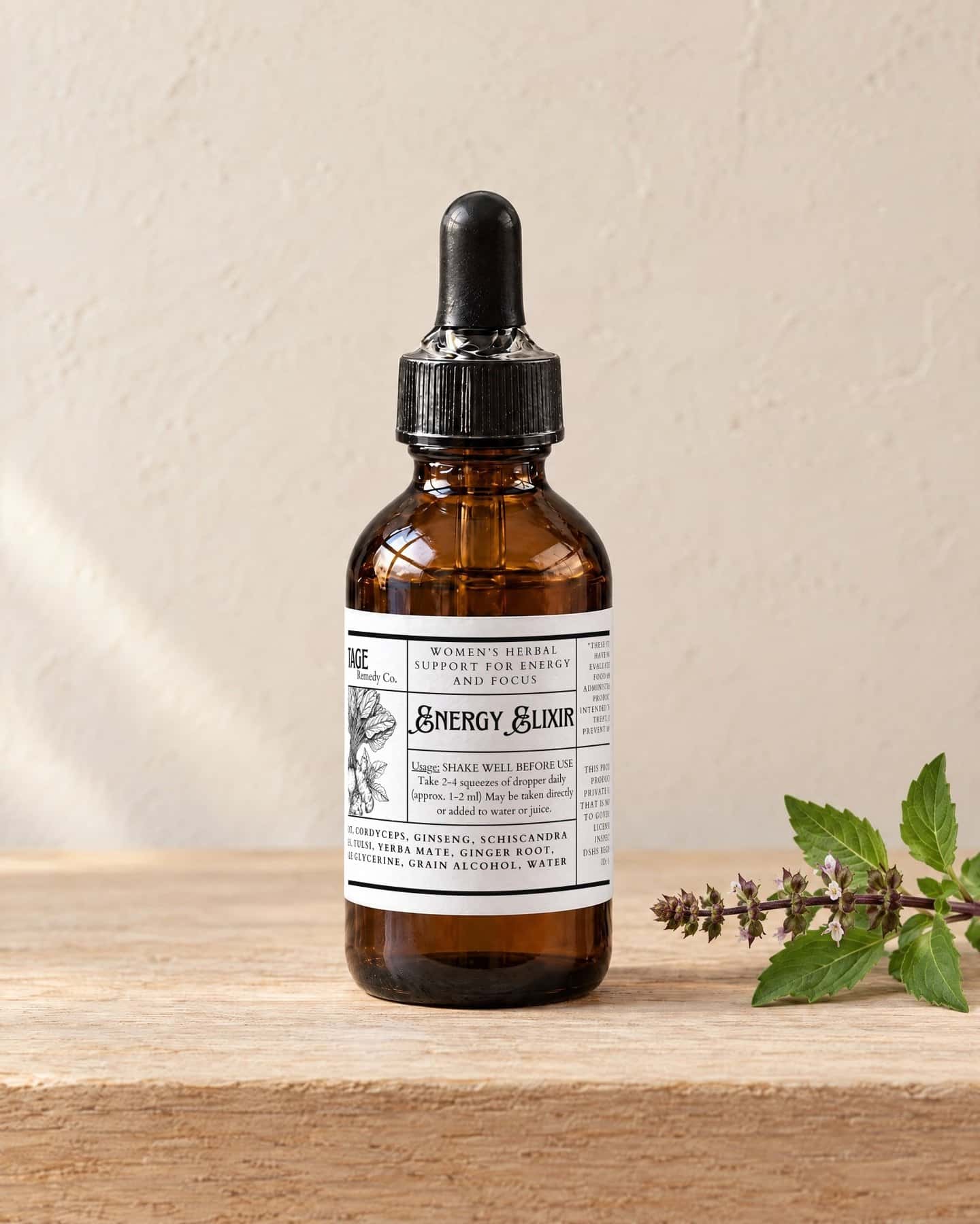A dropper bottle of EnergyElixir - Women's on a pale oak shelf, a sprig of fresh holy basil beside it