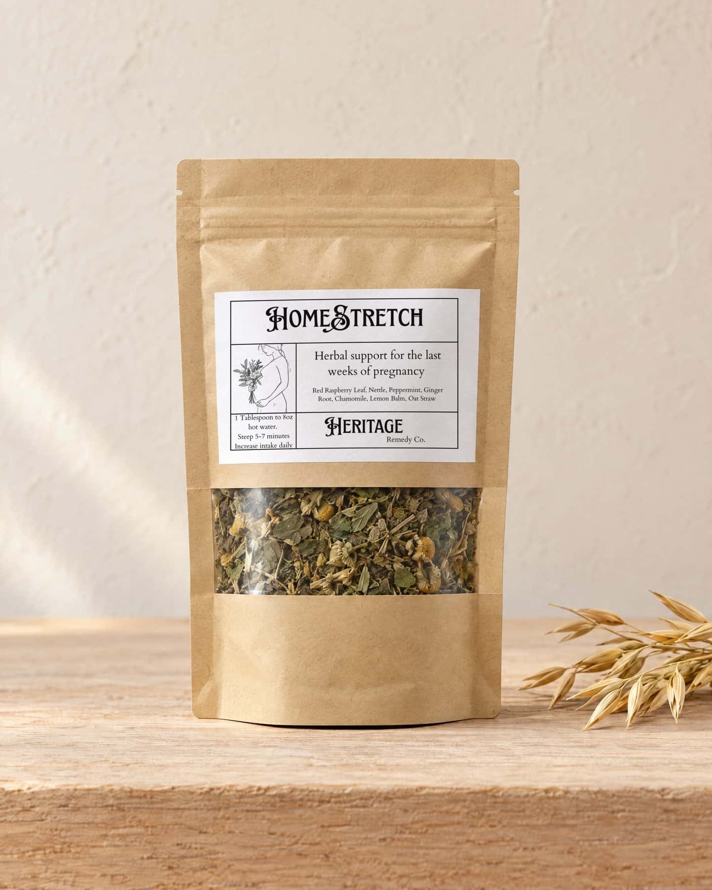 A kraft pouch of HomeStretch 3rd Trimester Tea with a window onto the loose tea, on a pale oak shelf, a few golden stalks of oat straw beside it