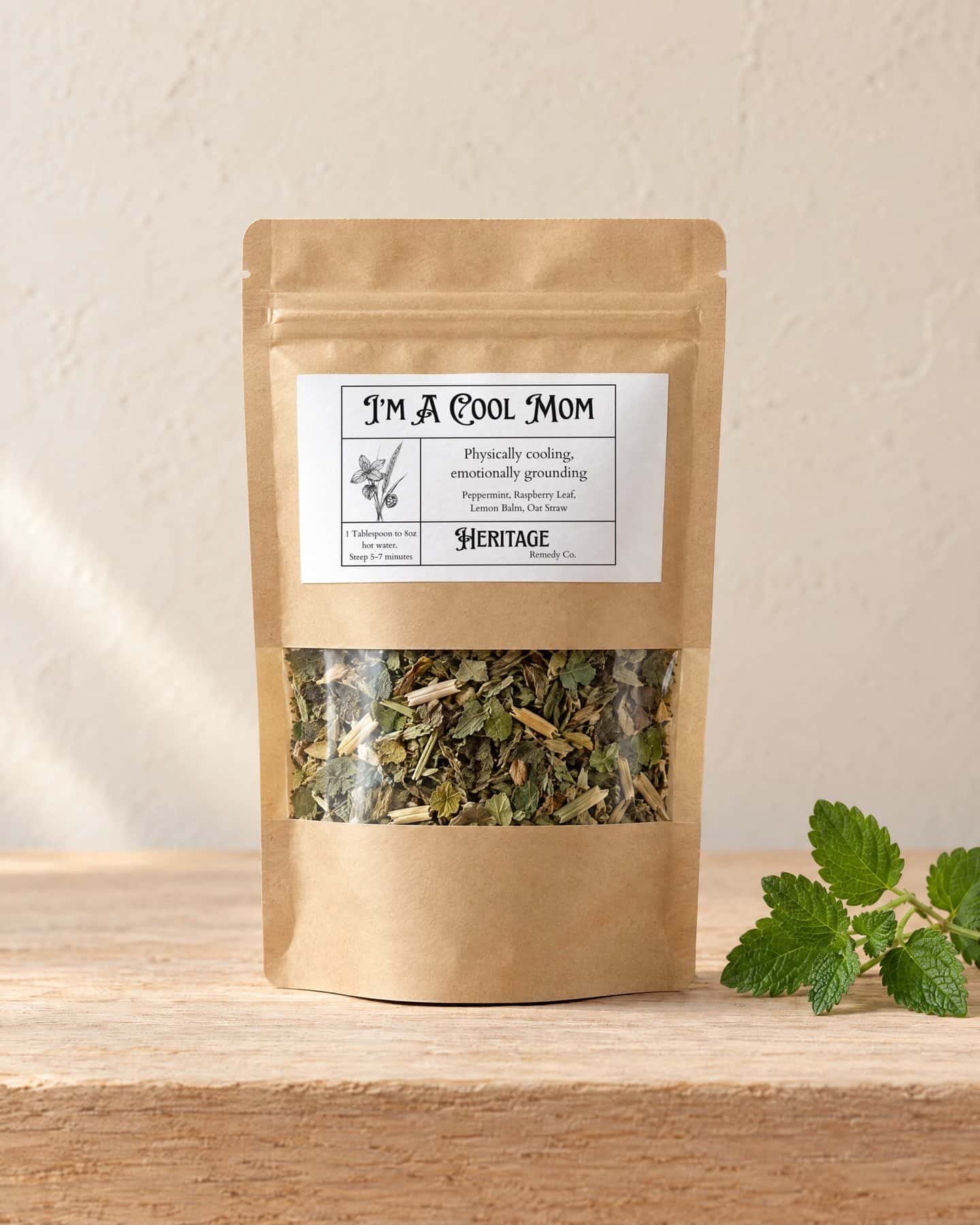 A kraft pouch of I'm A Cool Mom - Emotional Support with a window onto the loose tea, on a pale oak shelf, a sprig of fresh lemon balm beside it