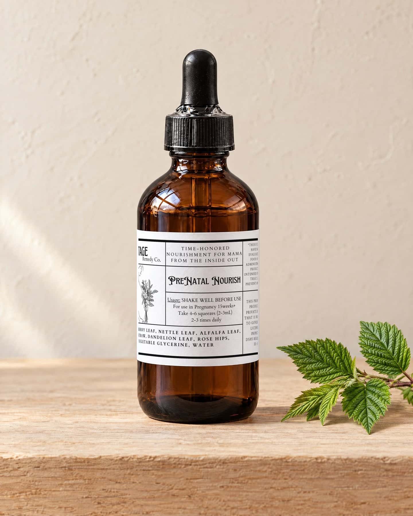 A four ounce dropper bottle of Prenatal Nourish on a pale oak shelf, a sprig of fresh red raspberry leaves beside it
